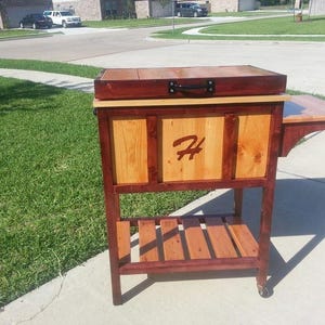 Könnte beinhalten: Ein Holz-Kühlschrank mit einer rot gebeizten Oberseite und einem Beistelltisch. Der Kühlschrank hat einen schwarzen Griff und ein geschnitztes "H" auf der Vorderseite. Der Kühlschrank steht auf Rädern.