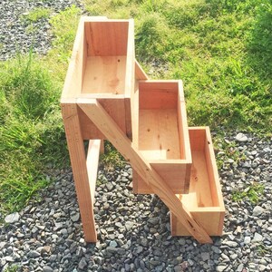 Peut inclure: Un bac &agrave; plantes en bois &agrave; trois niveaux avec un design ouvert. Le bac est construit en bois de couleur claire et comporte trois conteneurs rectangulaires de hauteurs variables, soutenus par des pieds inclin&eacute;s. La structure est pos&eacute;e sur un lit de gravier avec de l'herbe verte en arri&egrave;re-plan.
