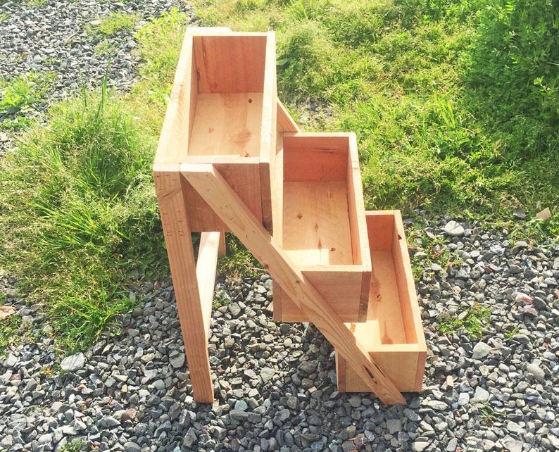 Peut inclure: Un bac &agrave; plantes en bois &agrave; trois niveaux avec un design ouvert. Le bac est construit en bois de couleur claire et comporte trois conteneurs rectangulaires de hauteurs variables, soutenus par des pieds inclin&eacute;s. La structure est pos&eacute;e sur un lit de gravier avec de l'herbe verte en arri&egrave;re-plan.