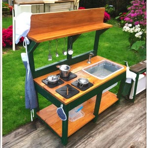 Puede incluir: Una cocina de juguete de madera con un marco pintado de verde y una encimera de madera marrón. La cocina tiene un fregadero, una estufa con cuatro quemadores y un estante para almacenamiento. También hay algunos utensilios de cocina colgados de ganchos en la parte posterior de la cocina.