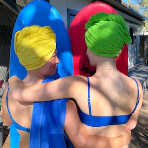 May include: Two people are standing next to each other, holding surfboards. One person is wearing a blue swimsuit and a green towel on their head. The other person is wearing a blue swimsuit and a yellow towel on their head. They are both smiling.