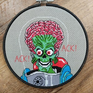 May include: A cross-stitch embroidery of a green alien with a pink brain, wearing a blue and gray spacesuit. The alien has red eyes and a wide, toothy grin. The text "ACK!" is stitched in red on the right side of the image.