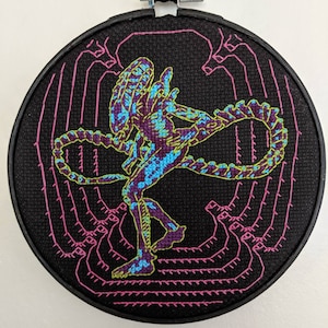 May include: A black embroidery hoop with a colorful embroidered image of an alien creature with a long tail. The alien is in a standing pose with its tail curled around its body. The background is black with pink and green stitching.
