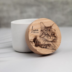 Puede incluir: Tapa de recuerdo para mascotas de madera con una imagen grabada de un gato. La tapa es redonda y presenta el nombre "Luna" y los años "2010-2025". La tapa está colocada sobre un recipiente cilíndrico blanco.