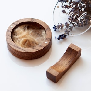 May include: A wooden keepsake box with a clear lid, engraved with the name "Jack" and the date "01-2023". The box contains a small amount of light brown hair. The box is sitting on a white surface with a small wooden stand next to it.