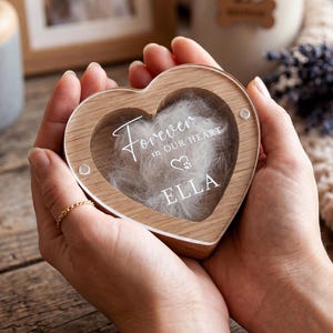 Op de afbeelding: Een hartvormig houten aandenkendoosje met een doorzichtig acryl voorpaneel. De tekst "Forever in our heart" staat in het wit gedrukt boven een klein hartje met een pootafdruk, gevolgd door de naam "ELLA" in het wit, gevuld met witte vacht.