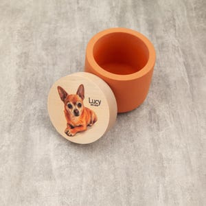 a wooden lid with a picture of a dog on it, and an orange ceramic container next to it.