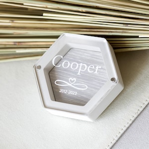 K&ouml;nnte beinhalten: Wei&szlig;e sechseckige Box mit einem durchsichtigen Deckel. Die Box hat ein graues Holzinnenleben mit dem Namen "Cooper" und den Daten "2012-2023" eingraviert. Die Box steht auf einer wei&szlig;en Oberfl&auml;che.