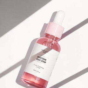 Pu&ograve; includere: Una bottiglia di vetro trasparente con un contagocce rosa riempita di un liquido rosa. La bottiglia ha un'etichetta bianca con il testo "BRIGHTENING BOOST SERUM" e "Vitamin C, Ginseng, Licorice" e "30ml / 1fl oz".