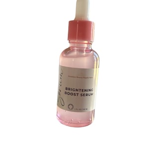 Pu&ograve; includere: Una bottiglia di vetro trasparente con un contagocce e un tappo rosa. La bottiglia contiene un siero etichettato "BRIGHTENING BOOST SERUM" e "1 FL OZ | 30 ML".