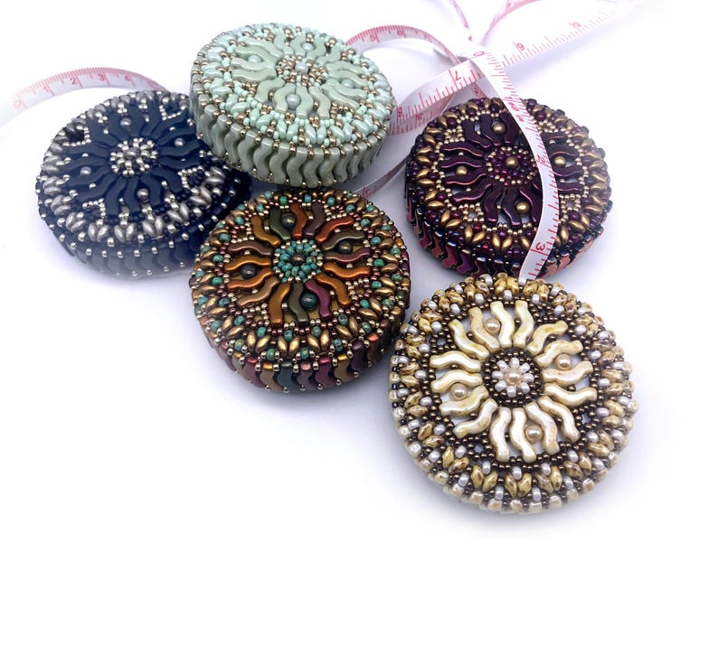 May include: Five beaded measuring tapes in different colors and patterns. The tapes are round and have a sunburst design. The colors include green, black, brown, purple, and white.