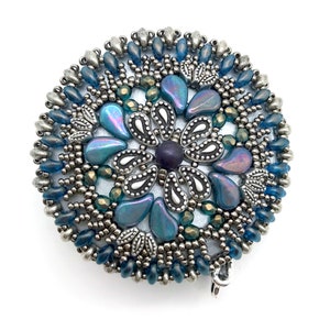 Puede incluir: Un broche redondo de cuentas plateadas y azules con un dise&ntilde;o de flor. El centro de la flor es una cuenta de color morado oscuro, y los p&eacute;talos est&aacute;n hechos de cuentas azules iridiscentes.
