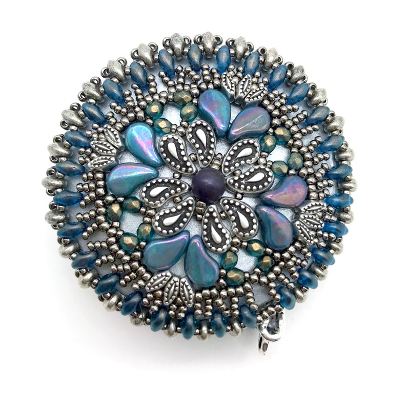 Puede incluir: Un broche redondo de cuentas plateadas y azules con un dise&ntilde;o de flor. El centro de la flor es una cuenta de color morado oscuro, y los p&eacute;talos est&aacute;n hechos de cuentas azules iridiscentes.