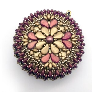 Puede incluir: Un colgante redondo de cuentas con un dise&ntilde;o de flor dorada y burdeos. El colgante est&aacute; hecho de peque&ntilde;as cuentas de semillas y tiene un acabado met&aacute;lico.