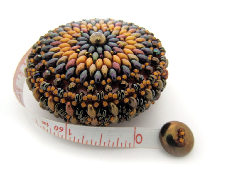 May include: A beaded measuring tape with a colorful, circular design. The tape is white with red markings and the numbers 1, 60, and 1 are visible.