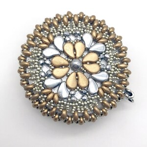 Puede incluir: Un broche redondo de cuentas doradas y plateadas con un dise&ntilde;o de flor. La flor est&aacute; hecha de cuentas plateadas y doradas, y el centro de la flor es una cuenta plateada.
