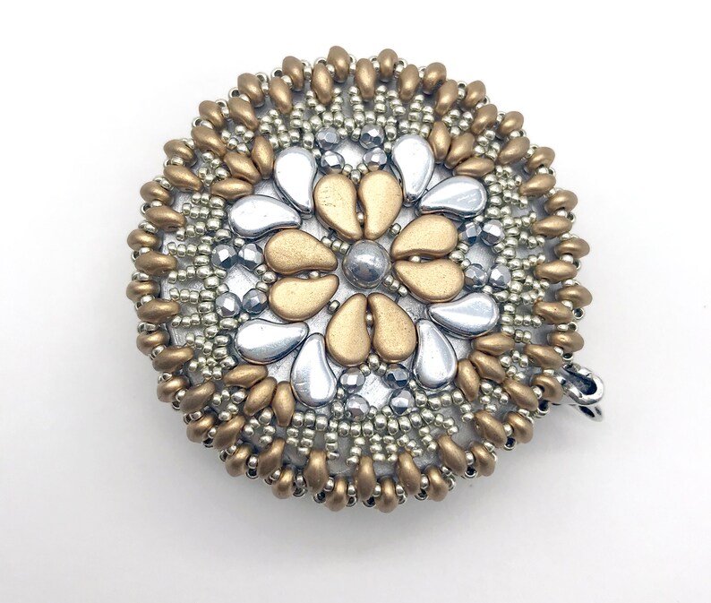 Puede incluir: Un broche redondo de cuentas doradas y plateadas con un dise&ntilde;o de flor. La flor est&aacute; hecha de cuentas plateadas y doradas, y el centro de la flor es una cuenta plateada.