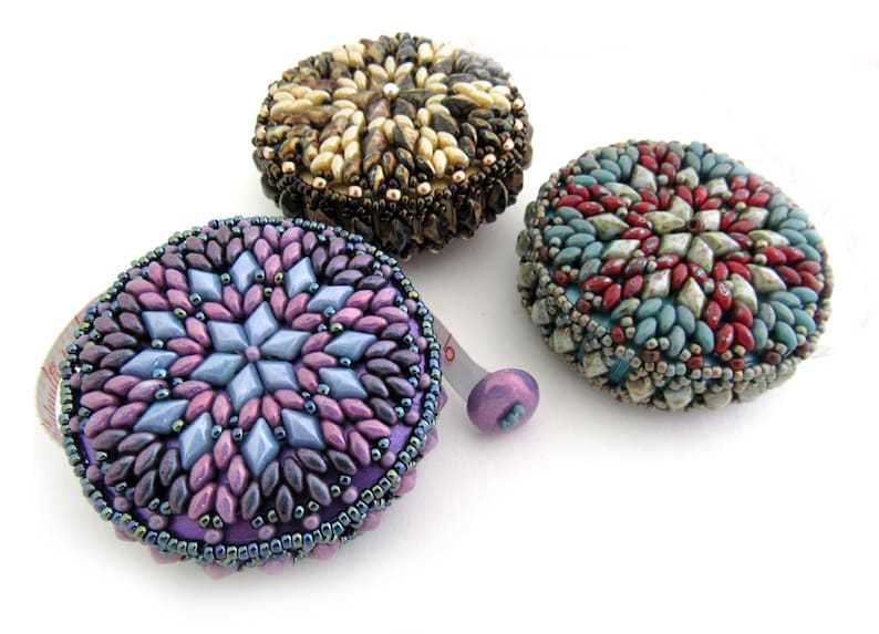 May include: Three beaded measuring tapes with different colored designs. The tape on the left has a purple, blue, and pink design. The tape in the middle has a brown, white, and gold design. The tape on the right has a red, blue, and green design.