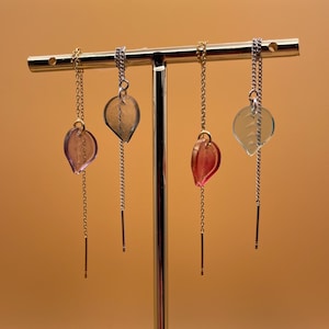 May include: Four pairs of dangle earrings with leaf-shaped charms. The charms are made of clear glass with a pink, red, or blue tint. The earrings are hanging from a silver stand.