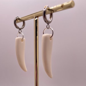 May include: A pair of white bone earrings with silver hoops and a gold stand. The earrings are hanging from the stand, and there are small stones on the surface below the stand.