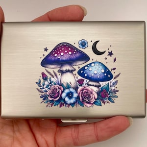 May include: A rectangular brushed silver metal card case featuring a colorful printed design of two mushrooms, one purple and one blue, surrounded by various flowers, leaves, small stars, and a crescent moon on the front surface of the case.