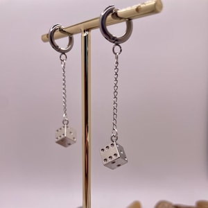 May include: A pair of silver hoop earrings with a dangling dice charm. The earrings are displayed on a wooden stand with a gold metal rod.