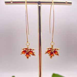 Peut inclure: Une paire de boucles d'oreilles pendantes dorées avec des motifs de feuilles d'érable en pierres précieuses rouges. Les boucles d'oreilles sont présentées sur un support en bois.
