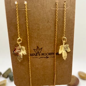 May include: Gold-colored threader earrings with maple leaf charms. The earrings are displayed on a brown card with the text "MAPLE & MOONSTONE jewelry". The leaves have intricate details and a reddish-brown hue.