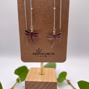 Peut inclure: Une paire de boucles d'oreilles en argent en forme de libellule avec une chaîne délicate. Les boucles d'oreilles sont présentées sur une carte brune avec le texte "Maple & Moonstone Jewelry".
