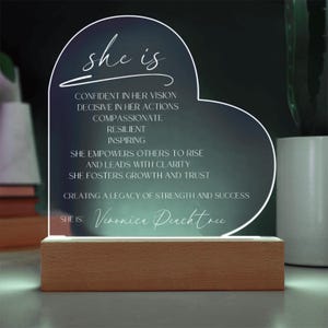 May include: A heart-shaped acrylic plaque with a white inscription that reads "she is" followed by a list of positive qualities. The inscription ends with "she is: Veronica Peachtree". The plaque is illuminated from behind and sits on a wooden base.