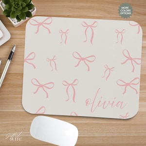 May include: A personalised mouse mat with a light beige background and a pattern of pink bows. The name "Olivia" is written in a cursive font. A computer mouse and pen are visible.