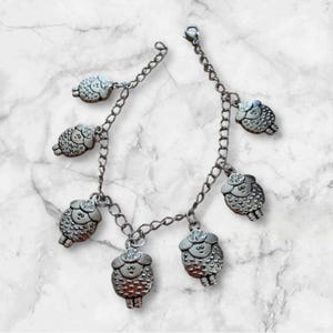 Puede incluir: Una pulsera de dijes plateada con múltiples dijes de oveja. La pulsera tiene un diseño de eslabones de cadena y los dijes de oveja están detallados con lana texturizada y rasgos faciales. La pulsera se exhibe sobre una superficie de mármol blanco y gris.