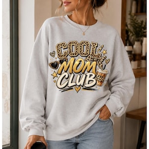 Peut inclure: Un sweat-shirt gris clair à col rond avec une impression graphique portant l'inscription COOL MOM CLUB en lettres stylisées. Le motif comprend des imprimés léopard, des cœurs, des étoiles, des lunettes, un nœud et une tasse de café.