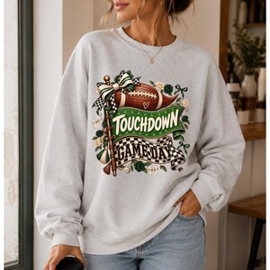 Peut inclure: Un sweat-shirt gris clair à col rond avec une impression graphique représentant un ballon de football marron, une bannière verte avec le texte "TOUCHDOWN" et une bannière à damier noir et blanc avec le texte "GAME DAY", orné de nœuds et de fleurs.