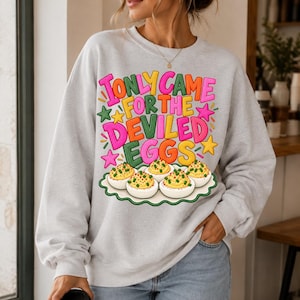 May include: A light gray crewneck sweatshirt featuring the colorful text I ONLY CAME FOR THE DEVILED EGGS in pink, yellow, and orange, accented by green and pink stars, and a graphic of a platter of five deviled eggs with green garnish.