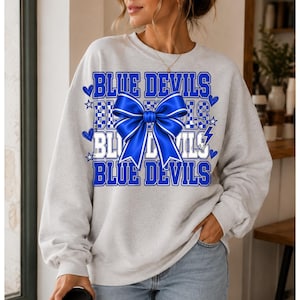 May include: A light gray crewneck sweatshirt featuring a graphic print with the text BLUE DEVILS repeated in blue and white block lettering, accented by a large, bright blue bow, small blue hearts, stars, and lightning bolt icons.