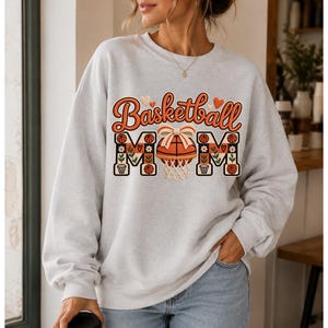 Peut inclure: Un sweat-shirt gris clair à col rond avec l'inscription Basketball Mom en orange et noir. Le motif comprend un ballon de basket avec un nœud, des petites fleurs et des cœurs. Le sweat-shirt a des manches longues et des bords-côtes.
