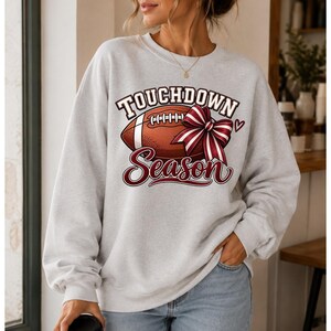 Peut inclure: Un sweat-shirt gris clair à col rond avec une impression graphique représentant un ballon de football américain marron à lacets blancs et un nœud rayé bordeaux et blanc. Le texte indique "TOUCHDOWN" en lettres blanches et "Season" en cursif.
