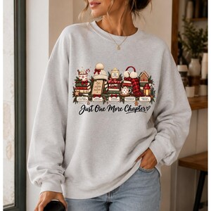 Peut inclure: Un sweat-shirt gris clair à col rond avec une impression graphique de livres empilés, une tasse, une bougie, un bonnet, des cache-oreilles et une maison en pain d'épices. Le texte "Just One More Chapter" est imprimé en noir sous l'illustration.