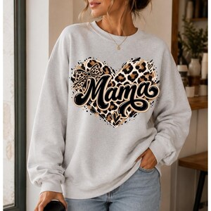 Peut inclure: Un sweat-shirt gris clair à col rond avec un grand motif en forme de cœur sur la poitrine. Le cœur présente un imprimé léopard avec un nœud noir et le mot Mama écrit dans une police cursive noire et grasse avec un contour blanc.