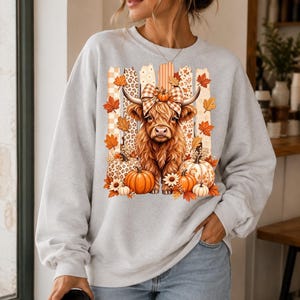 Peut inclure: Un sweat-shirt gris clair à col rond avec une impression graphique représentant une vache Highland brune et poilue portant un nœud à carreaux. Elle est entourée de citrouilles orange, de feuilles d'automne et de panneaux verticaux.