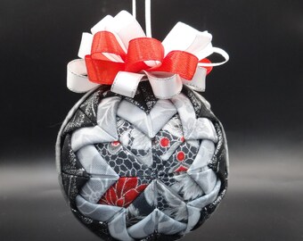 May include: A spherical fabric ornament featuring a folded patchwork design in black, white, and red floral and lace patterns. The ornament is topped with a decorative bow made of layered white and red ribbons and hangs from a thin white cord.