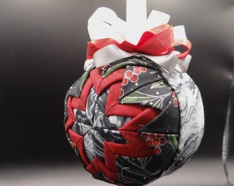 May include: A handmade Christmas ornament with a patchwork design. The ornament features red, black, and white fabric pieces arranged in a geometric pattern. A white ribbon and red ribbon bow are at the top, with a white string for hanging.
