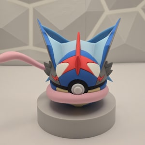 Könnte beinhalten: Detaillierte Figur einer blau, rot und schwarz gefärbten Figur mit rosa Zunge, auf einem grauen Sockel. Die Figur hat große Augen und eine markante Kopfbedeckung. Der Hintergrund zeigt ein geometrisches Muster.