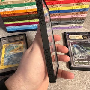 Pu&ograve; includere: Custodia protettiva nera con la scritta "SLABSTAR" e "GEOFARM" sul lato, che contiene una carta collezionabile valutata. Sullo sfondo, pile di scatole colorate per la conservazione delle carte, ciascuna con l'etichetta "SLABSTAR" e "GEOFARM".
