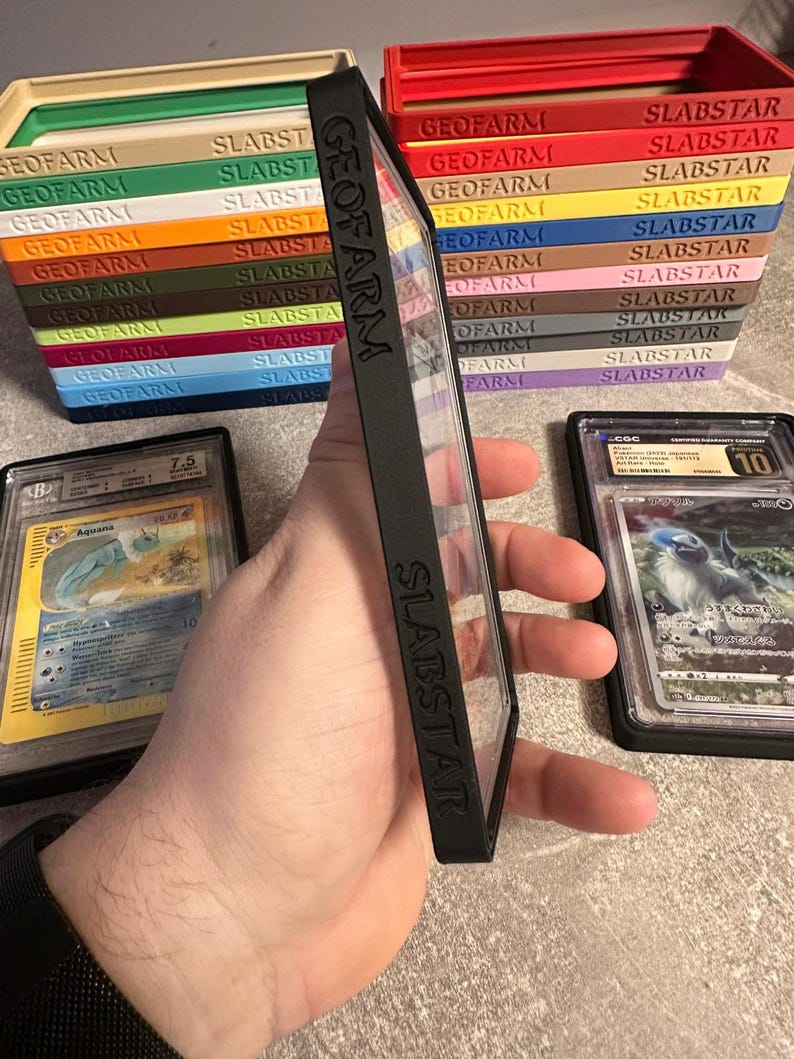 Pu&ograve; includere: Custodia protettiva nera con la scritta "SLABSTAR" e "GEOFARM" sul lato, che contiene una carta collezionabile valutata. Sullo sfondo, pile di scatole colorate per la conservazione delle carte, ciascuna con l'etichetta "SLABSTAR" e "GEOFARM".