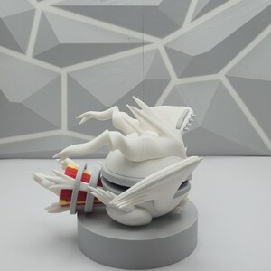 May include: A white figurine featuring a spherical body with wing-like appendages and a tail-like structure. The tail section displays red and yellow accents. The figure is positioned on a circular gray base, showcasing a smooth, matte finish.