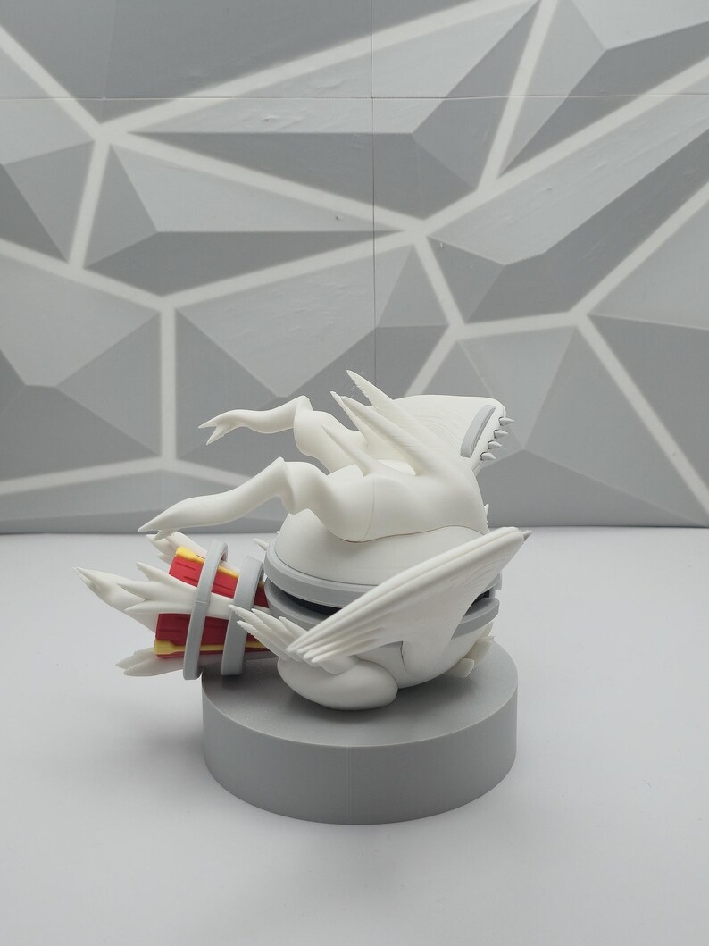 May include: A white figurine featuring a spherical body with wing-like appendages and a tail-like structure. The tail section displays red and yellow accents. The figure is positioned on a circular gray base, showcasing a smooth, matte finish.