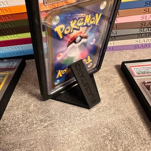 Pu&ograve; includere: Un espositore in plastica nera che tiene una carta Pok&eacute;mon valutata. La carta &egrave; racchiusa in un supporto di plastica trasparente con il logo Pok&eacute;mon visibile. Il supporto &egrave; etichettato "SLABSTAND" ed &egrave; su una superficie strutturata.