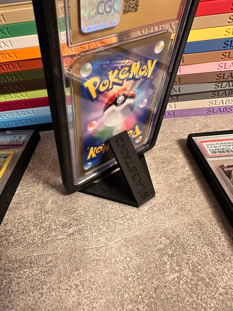 Pu&ograve; includere: Un espositore in plastica nera che tiene una carta Pok&eacute;mon valutata. La carta &egrave; racchiusa in un supporto di plastica trasparente con il logo Pok&eacute;mon visibile. Il supporto &egrave; etichettato "SLABSTAND" ed &egrave; su una superficie strutturata.